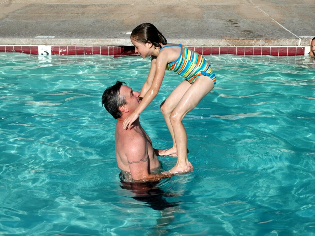 Un padre que se pregunta qué hacer si mi hijo no quiere meterse al agua, y ayudando a su hija a meterse a una alberca, jugando